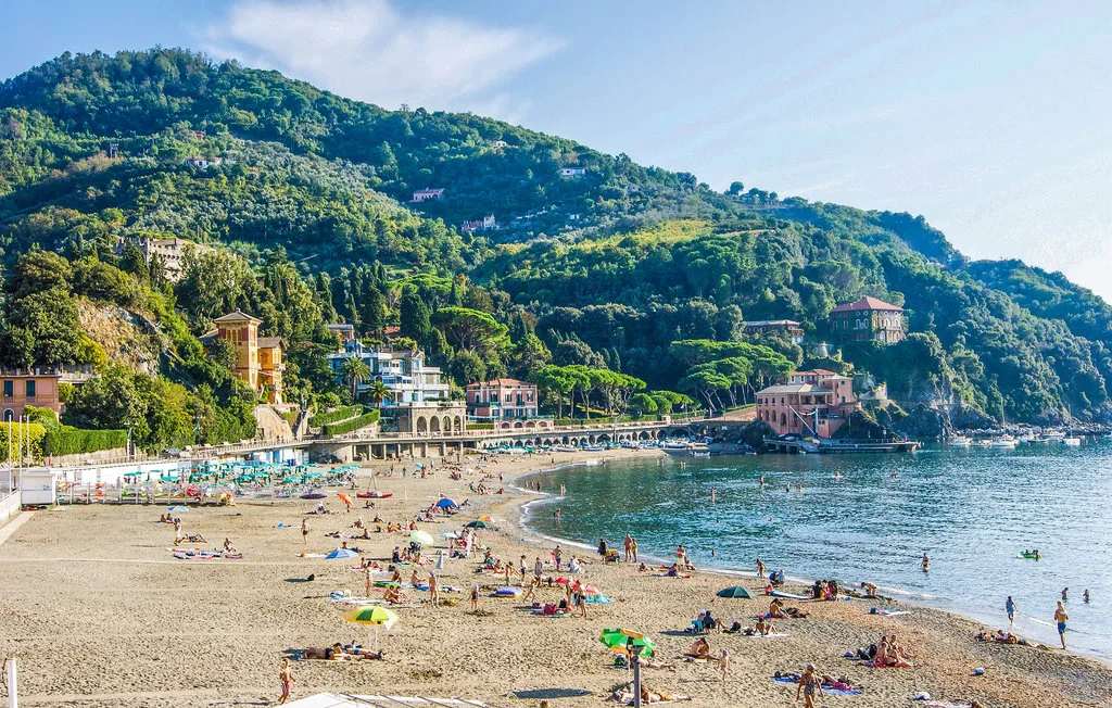 A coastal beach with people on the sand and forested hills behind it.