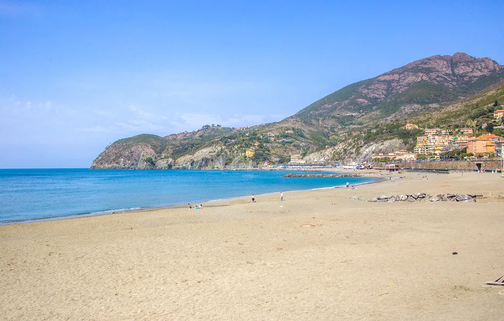 A sandy beach beside calm blue water, with a rocky coastline and hills in the distance.