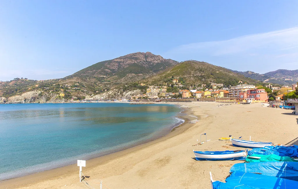 A sandy beach curves around a calm bay, with boats along the shore.