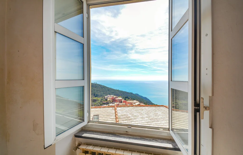 An open window overlooks rooftops, a green hillside, and the sea.