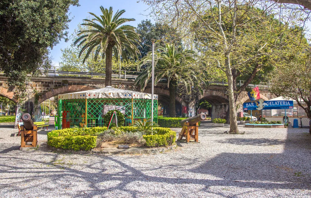 A tree-shaded plaza with a small kiosk and a stone bridge.