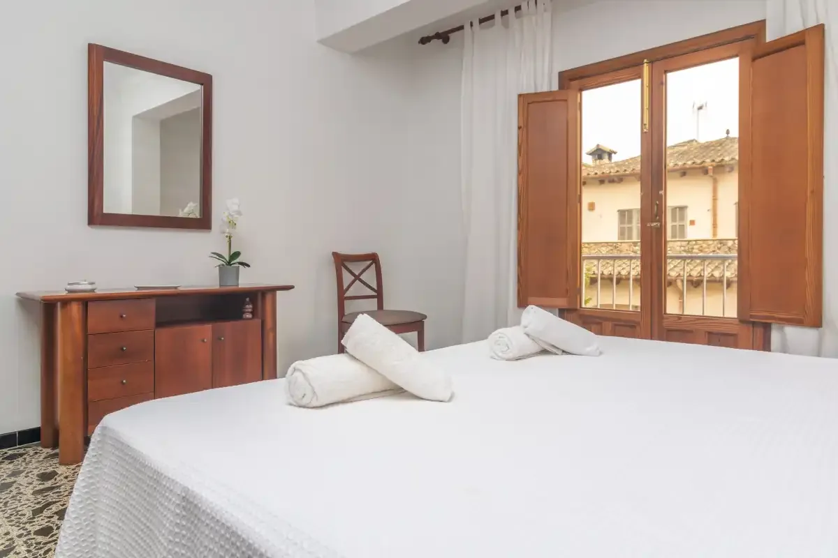 A bedroom with a neatly made bed and open wooden shutters overlooking a balcony.