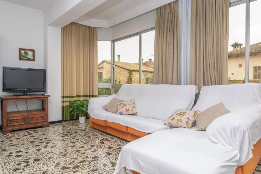 A bright living room with white-covered seating beside large windows and beige curtains.
