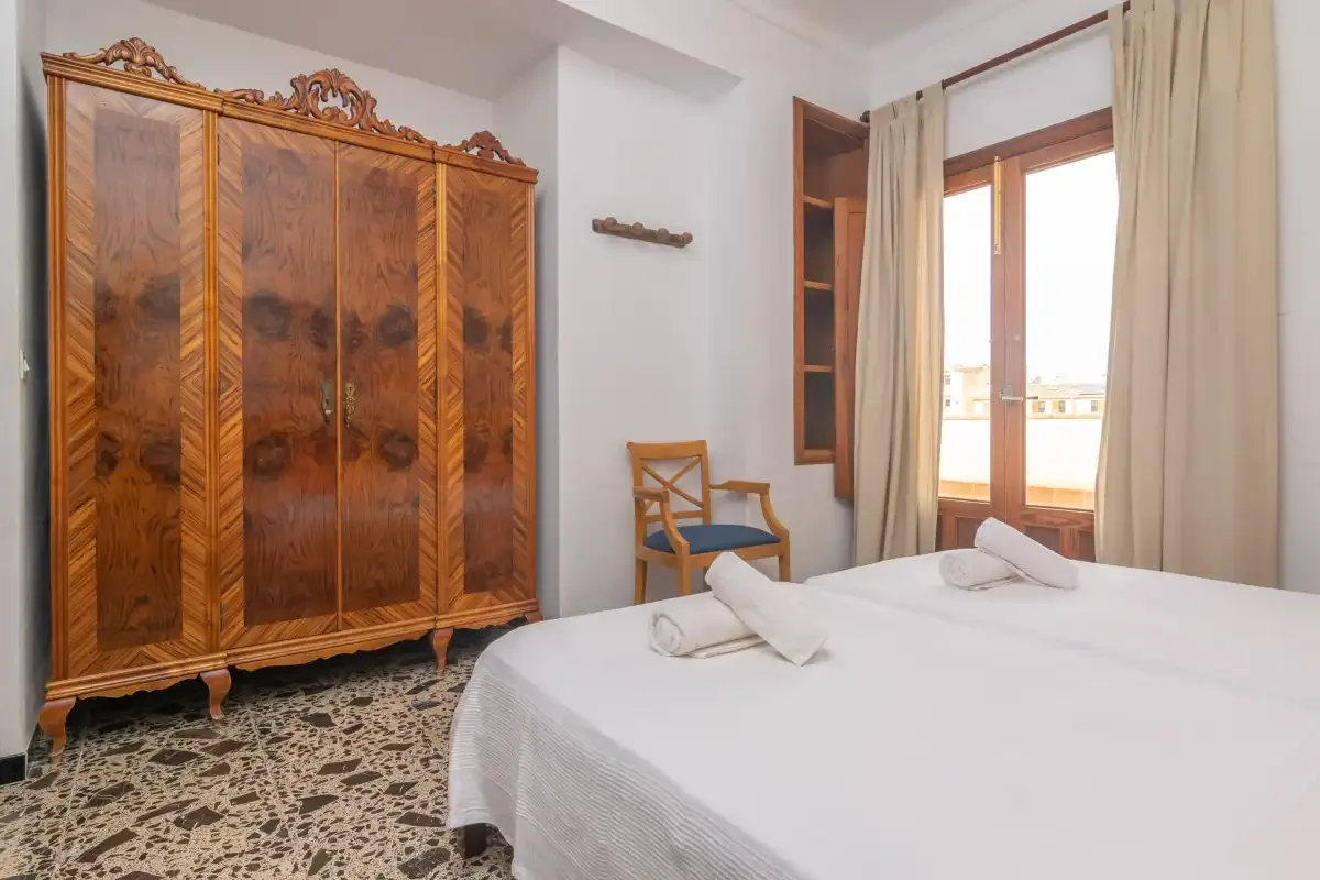 A bedroom with a large wooden wardrobe and a bed beside a curtained window.