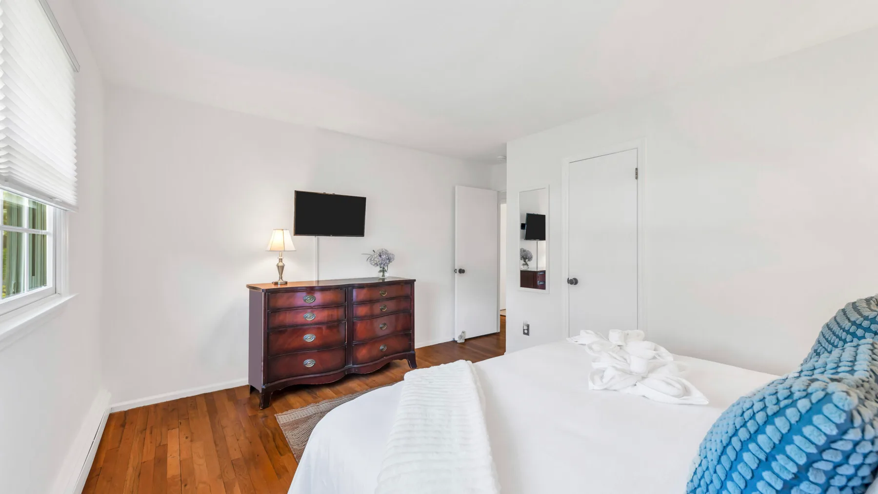 A bright bedroom with a bed, wooden dresser, and wall-mounted television.