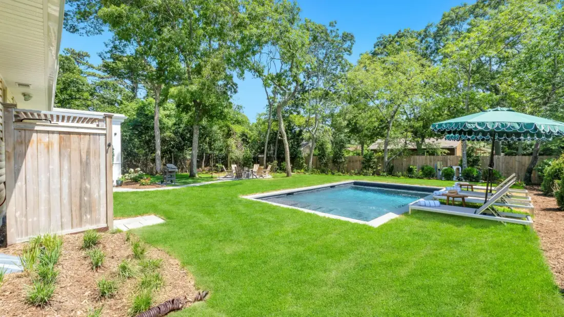 A landscaped backyard features a swimming pool, green lawn, and shaded seating area.