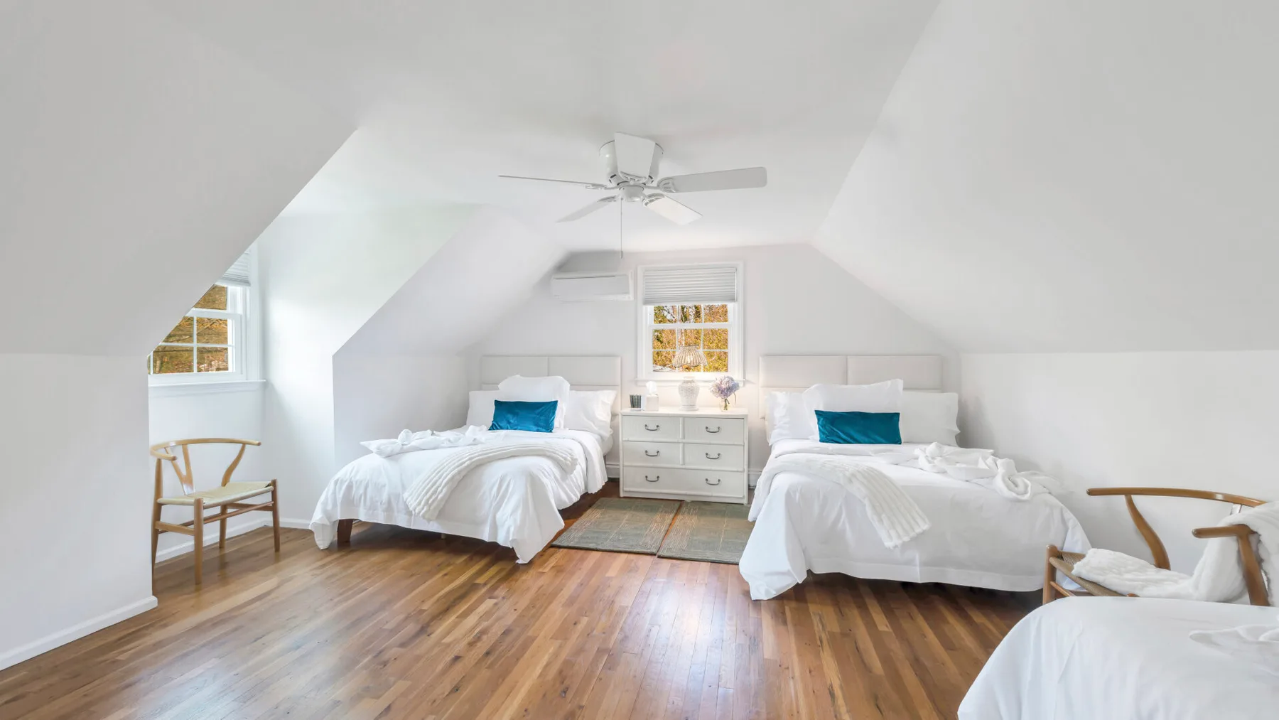 A bright bedroom with white bedding, wooden flooring, and a ceiling fan.