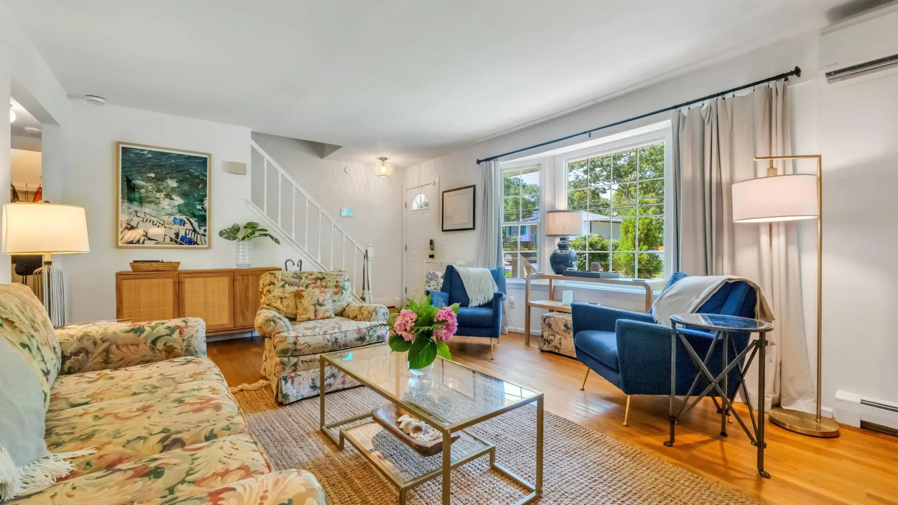 A bright living room with floral seating and large windows overlooking greenery.