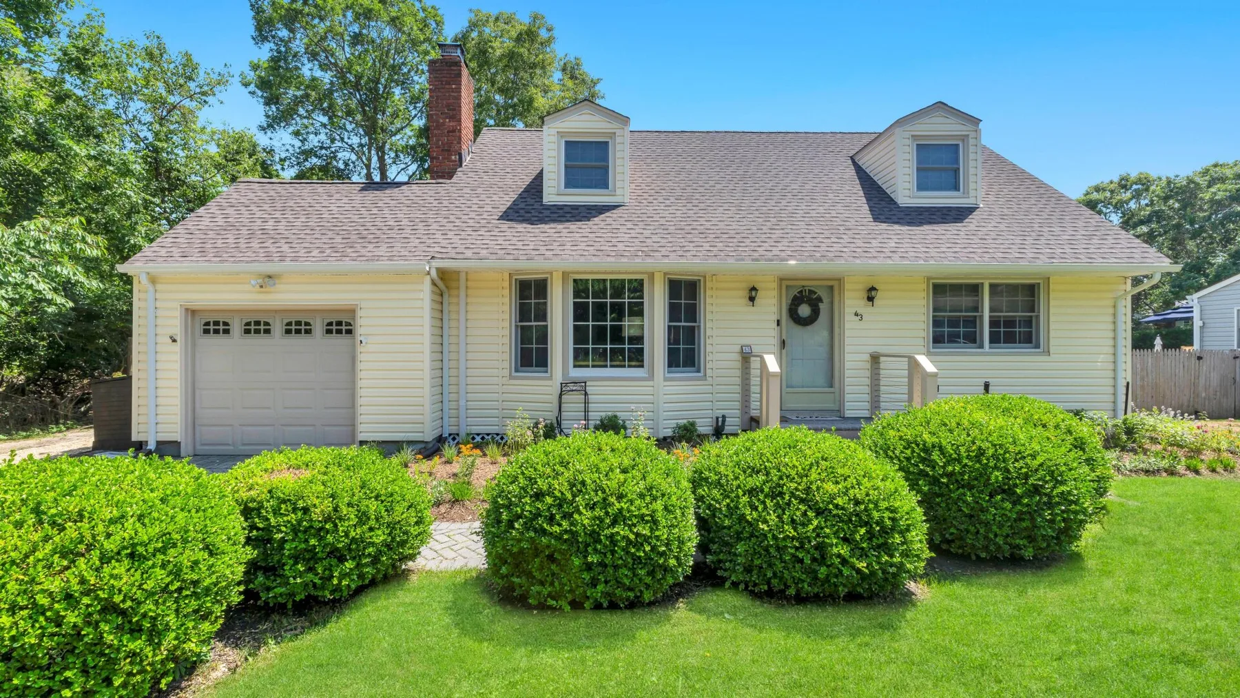 A light-colored house with a garage sits behind a lawn and trimmed shrubs.
