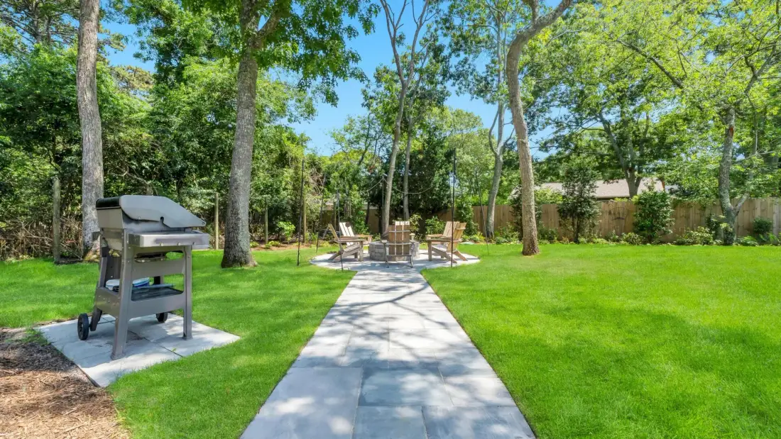 A paved garden path leads through a green lawn toward outdoor seating beneath trees.