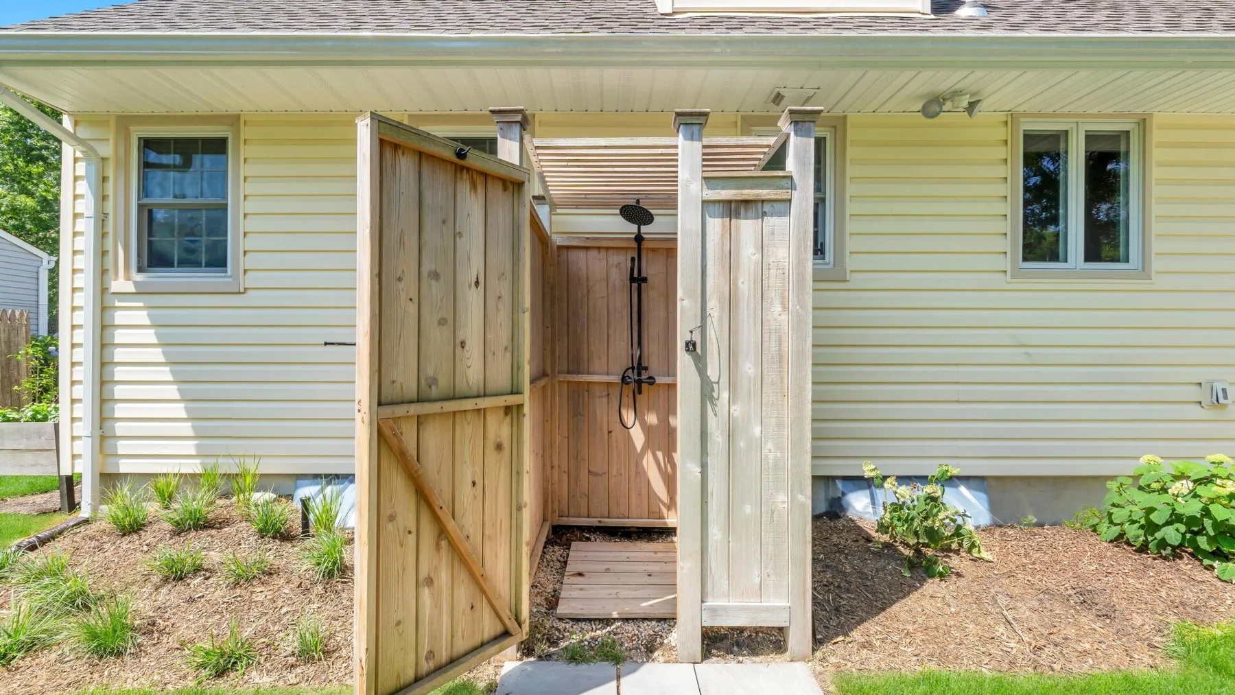 An outdoor shower is screened by wooden panels beside a light-colored house.