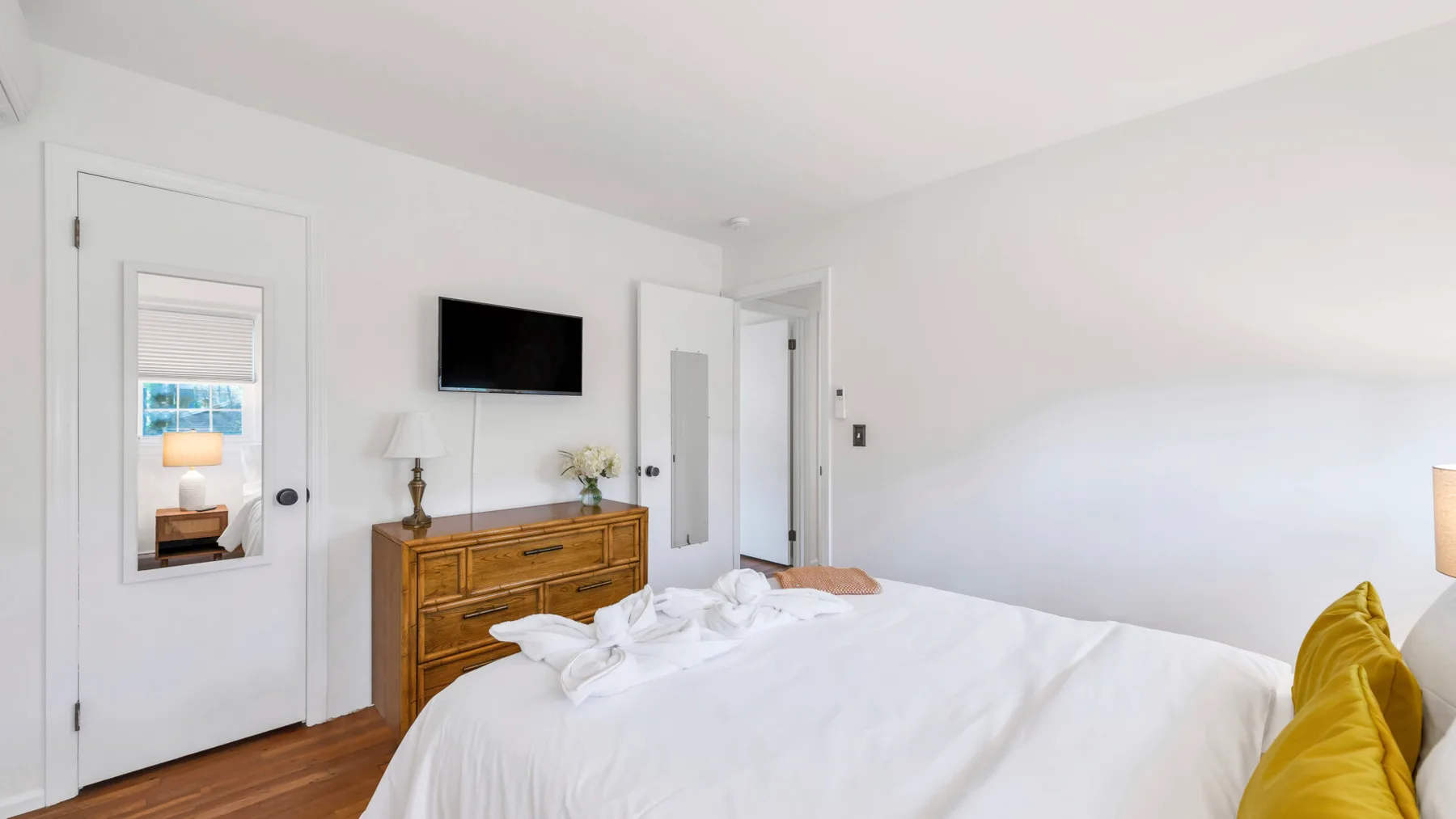 A bright bedroom with a white bed, wooden dresser, and wall-mounted television.