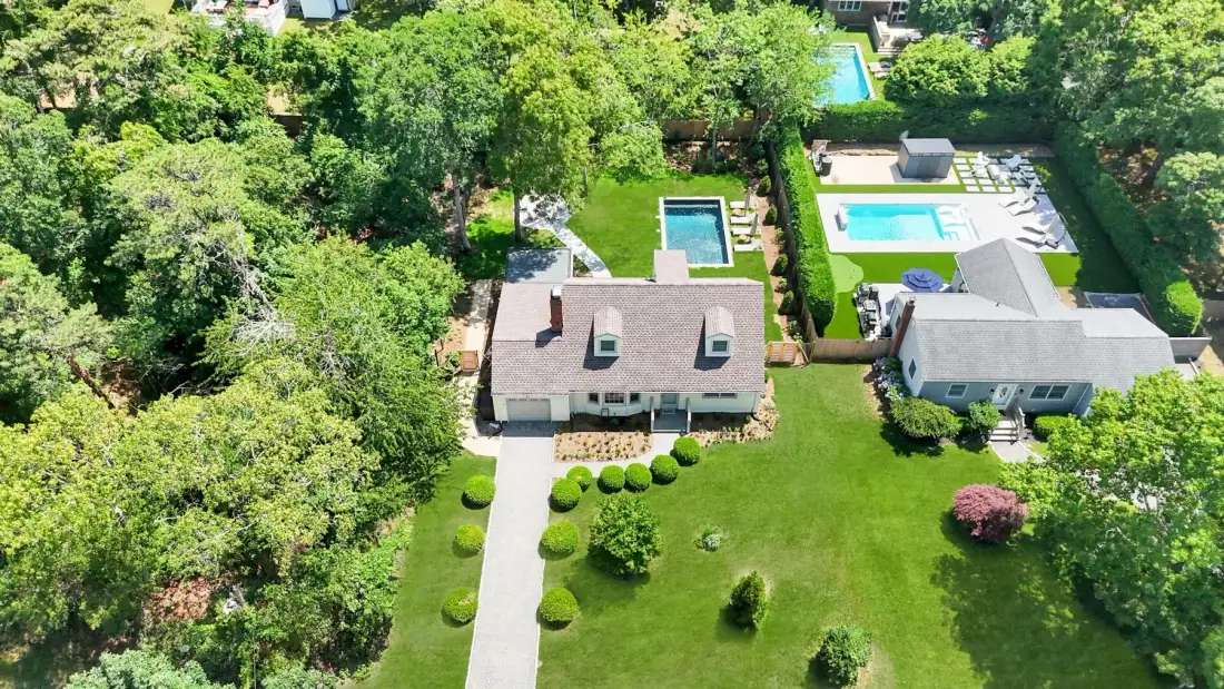 An aerial view shows two houses, green lawns, and several swimming pools among trees.