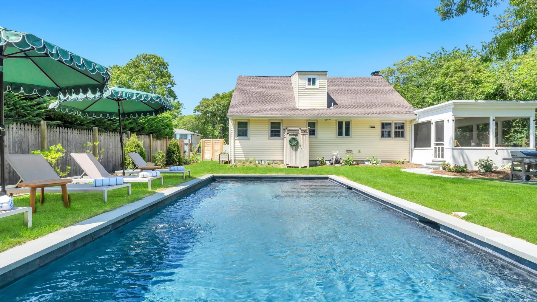 Una piscina exterior junto a una casa, con asientos y césped verde.