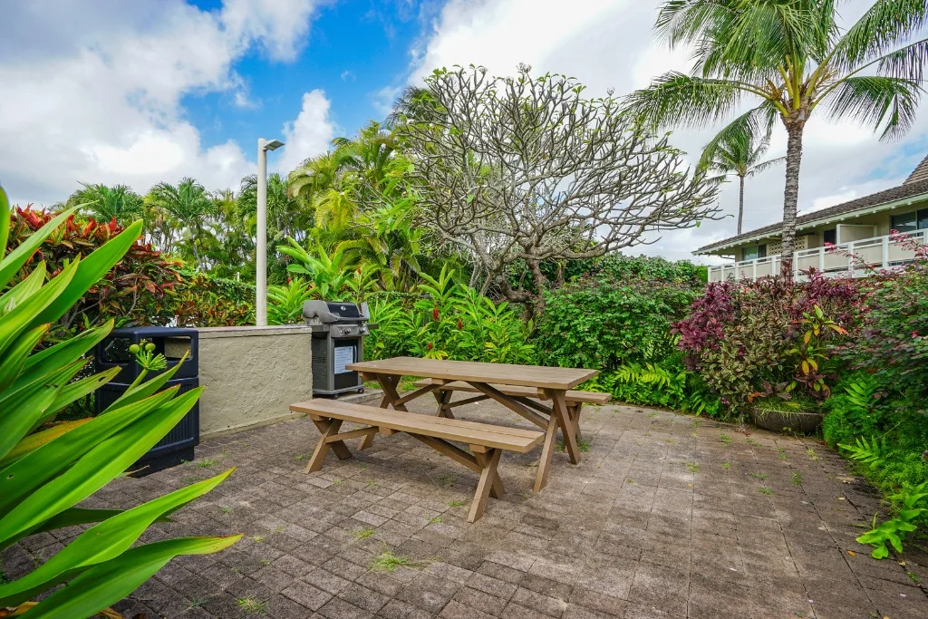 Un patio exterior tiene una mesa de picnic de madera rodeada de vegetación tropical.