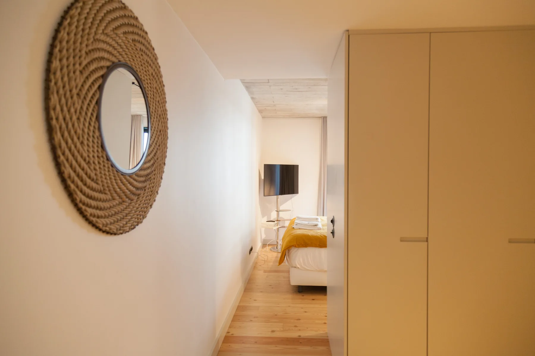 Se ve un dormitorio al fondo de un pasillo estrecho, con un espejo redondo en la pared.