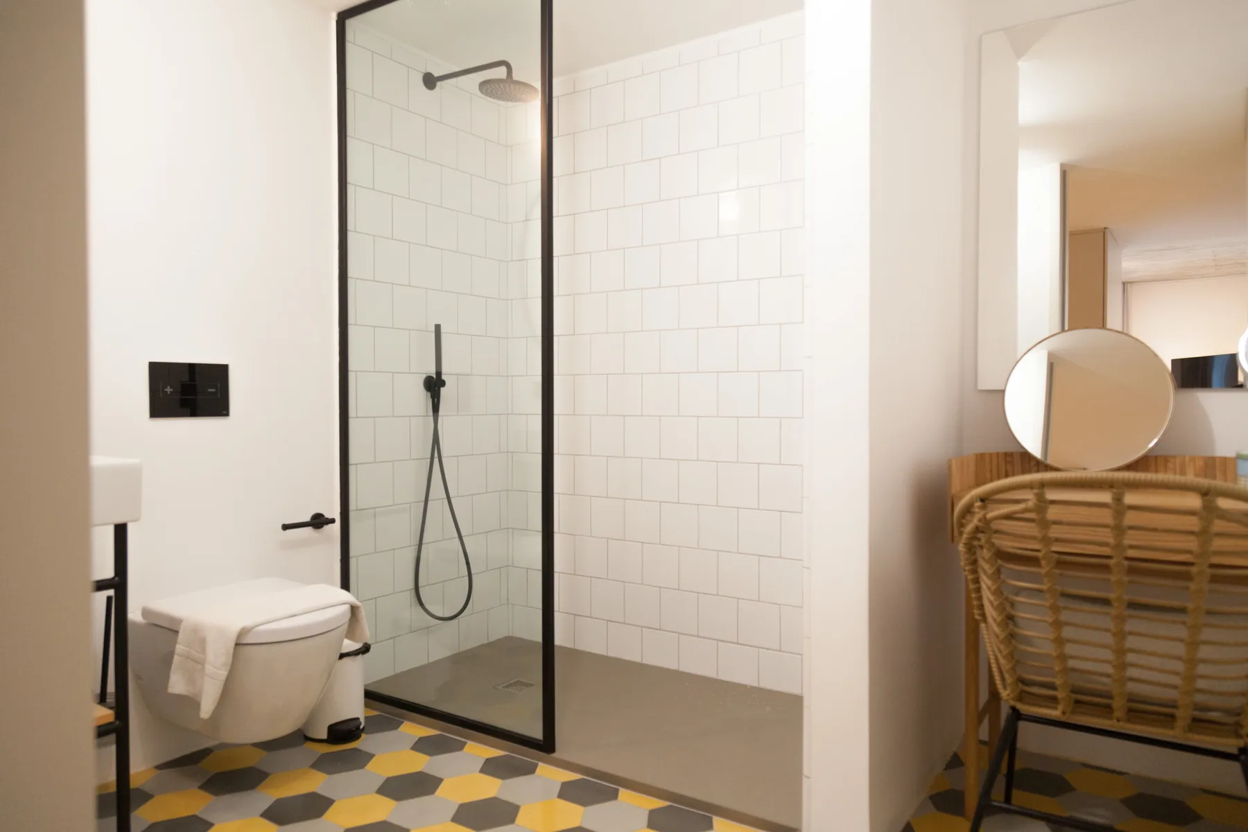 Un baño luminoso tiene una ducha de vidrio, un inodoro suspendido y azulejos geométricos en el suelo.