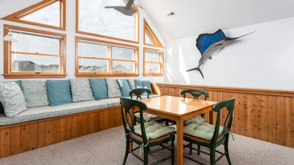 Comedor con una mesa de madera y un banco acolchado bajo ventanas grandes.