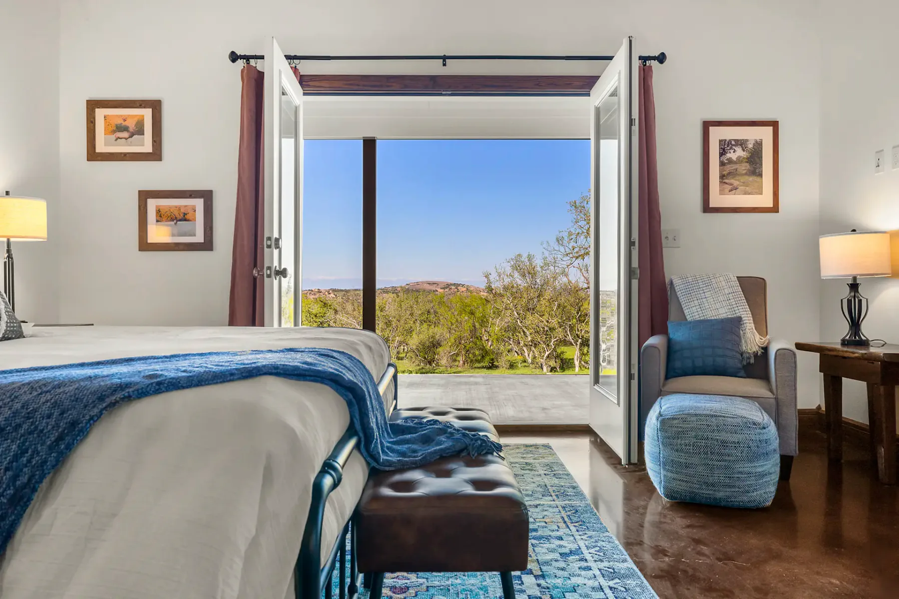 Dormitorio luminoso con cama, manta azul, reposapiés y sillón. Las puertas abiertas dejan ver un paisaje pintoresco. La iluminación cálida y las obras de arte enmarcadas adornan las paredes.