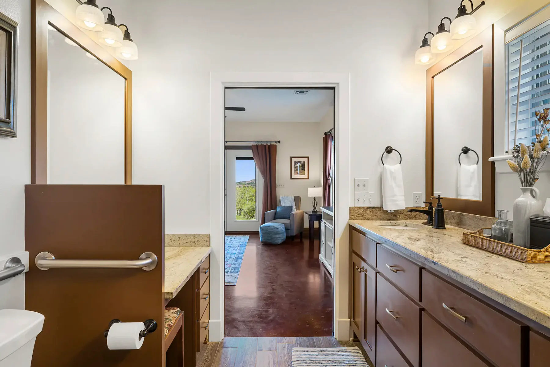 Un baño con un tocador largo y un espejo da paso a un dormitorio con ventana.