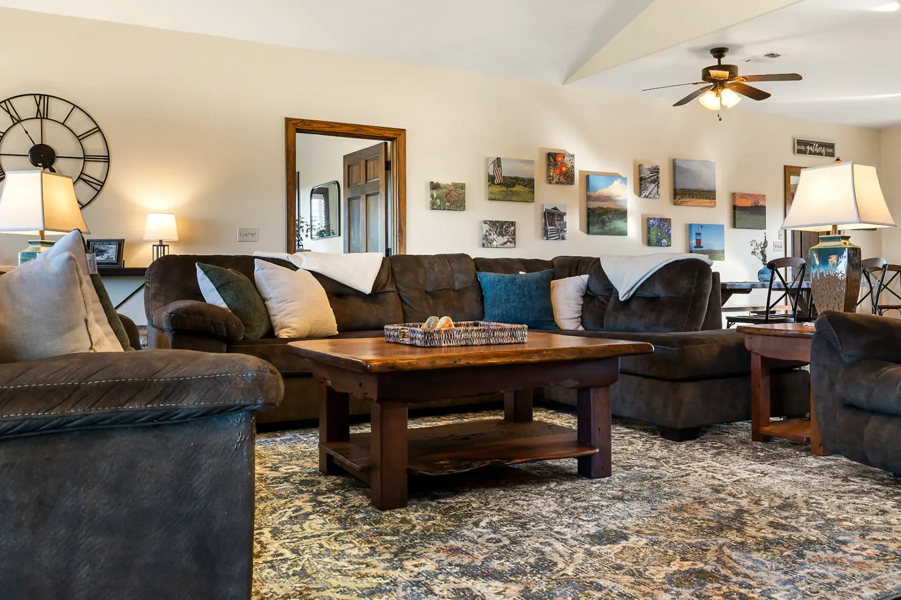 Cozy living room with dark sofas, a wooden coffee table, patterned rug, wall art, a large clock, and ceiling fan.