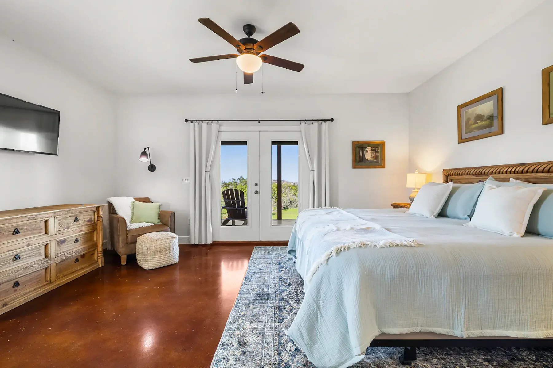Dormitorio luminoso con cama grande, ventilador de techo, cuadros, cómoda, televisor y sillón. Unas puertas francesas dan a un patio con sillas de exterior.