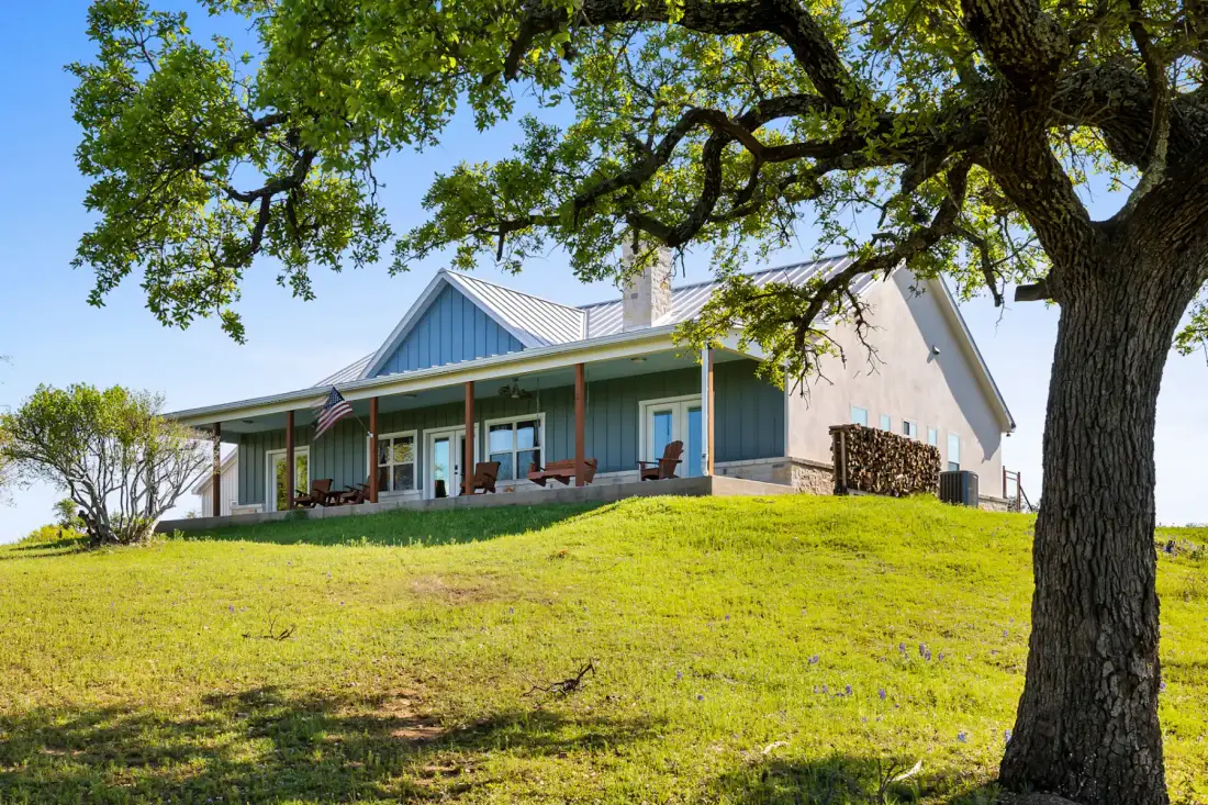 Una casa moderna de estilo rancho con porche, rodeada de césped verde y árboles, bajo un cielo azul despejado.