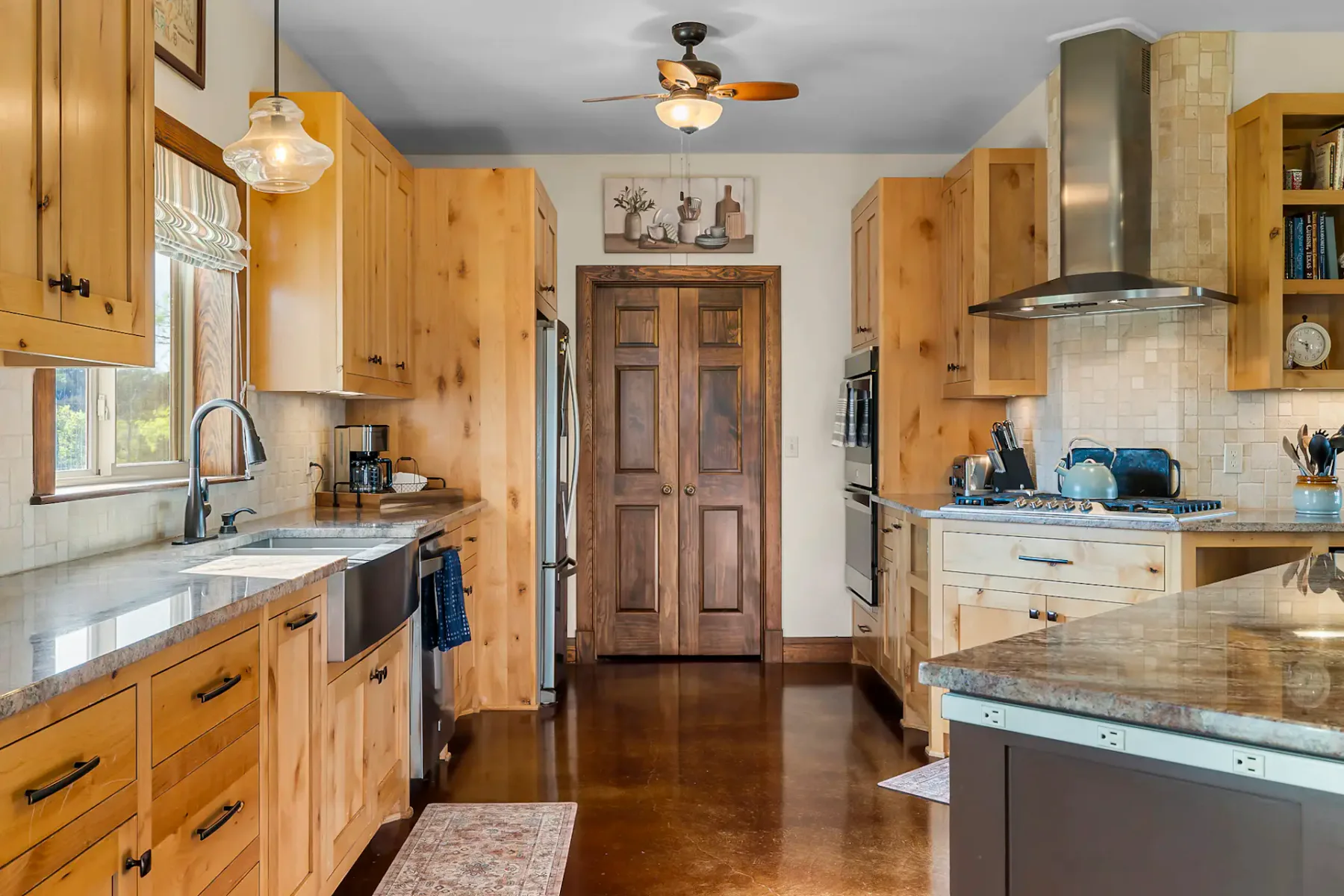 Cocina cálida y rústica con armarios de madera, encimeras de piedra, electrodomésticos de acero inoxidable y ventilador de techo. La luz entra por una ventana.