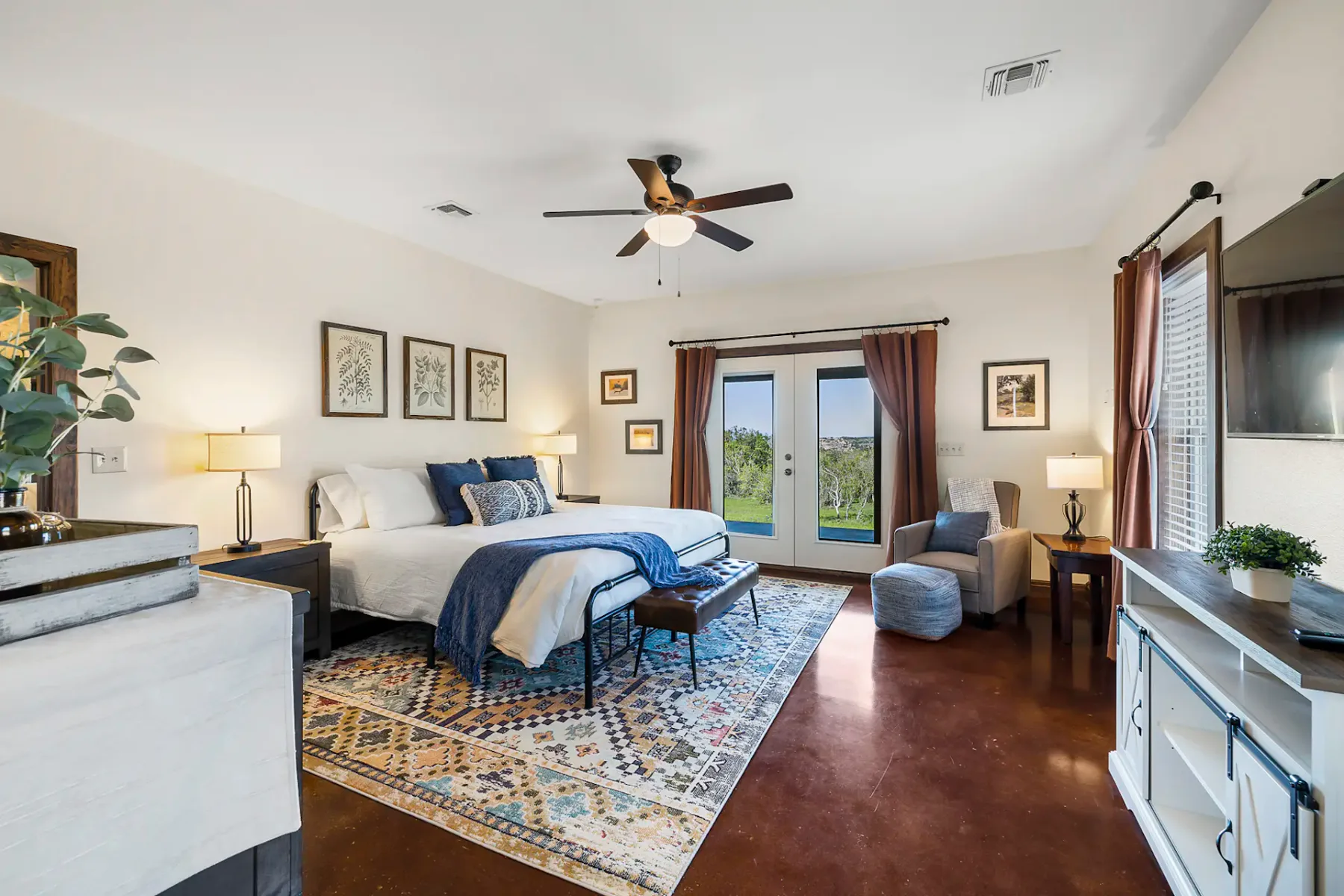 Dormitorio con cama grande, ropa de cama azul, alfombra estampada, ventilador de techo, cuadros enmarcados y vistas a través de unas puertas dobles. Acogedora zona de estar junto a la ventana.
