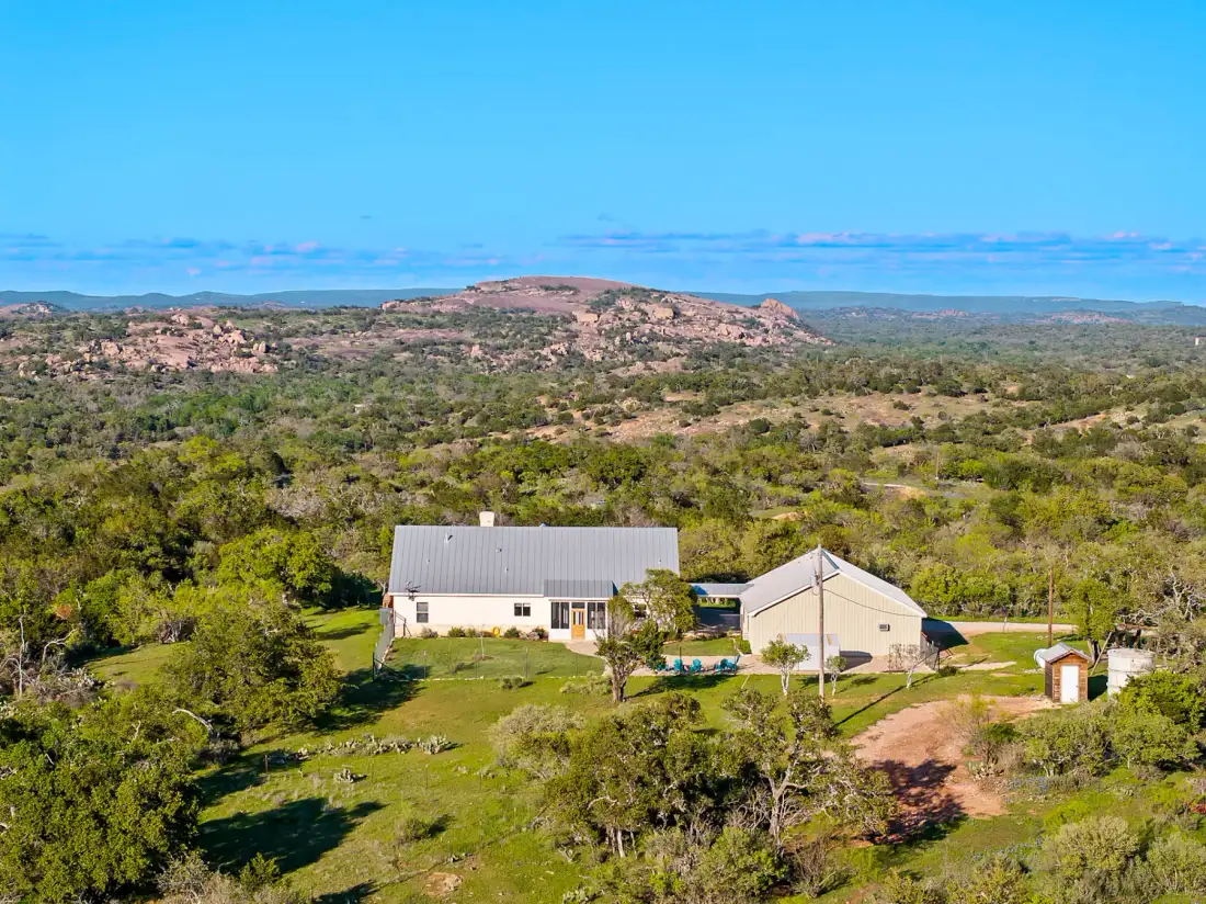 Vista aérea de una casa rural y un granero rodeados de extensas zonas verdes y colinas bajo un cielo azul despejado.