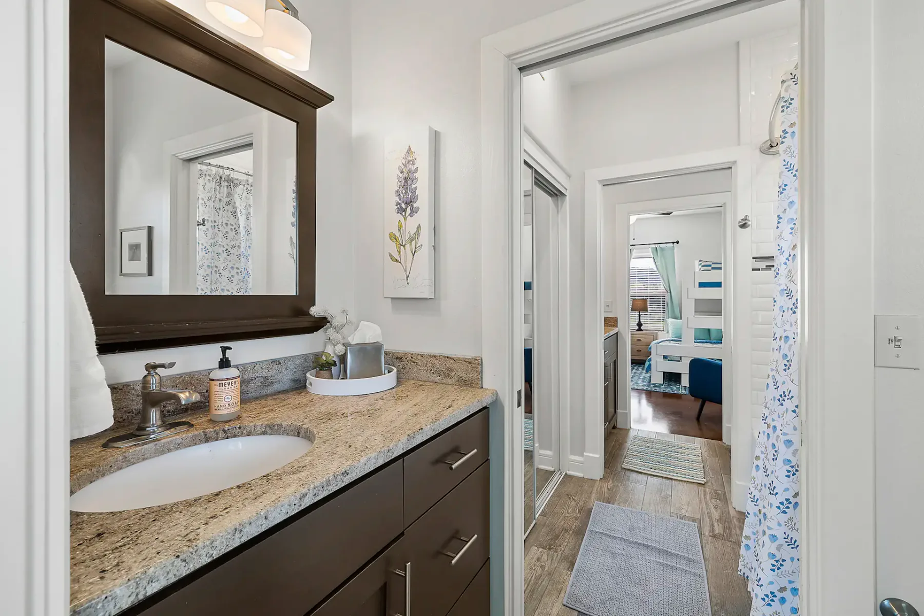 Baño moderno con encimera de granito, mueble de lavabo de madera oscura y espejo. La vista se extiende hacia un pasillo y una habitación al fondo con decoración azul y blanca.