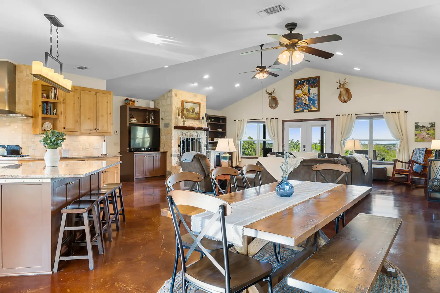 Spacious open-plan living area with dining table, kitchen island, wooden cabinets, ceiling fans, and large windows with scenic views.