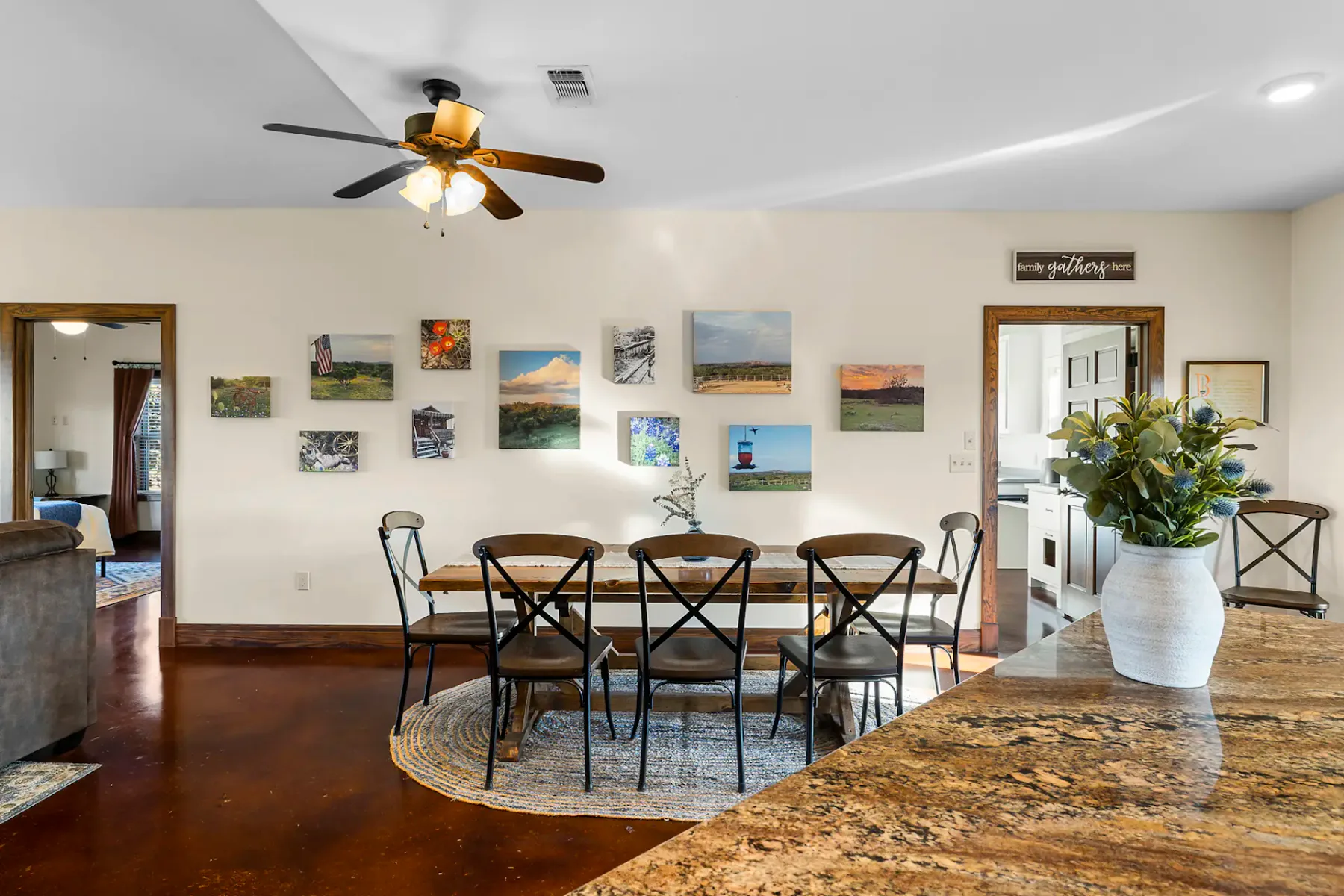 Dining area with a wooden table and eight chairs, wall art, ceiling fan, and a view into another room. A vase with flowers on the counter.