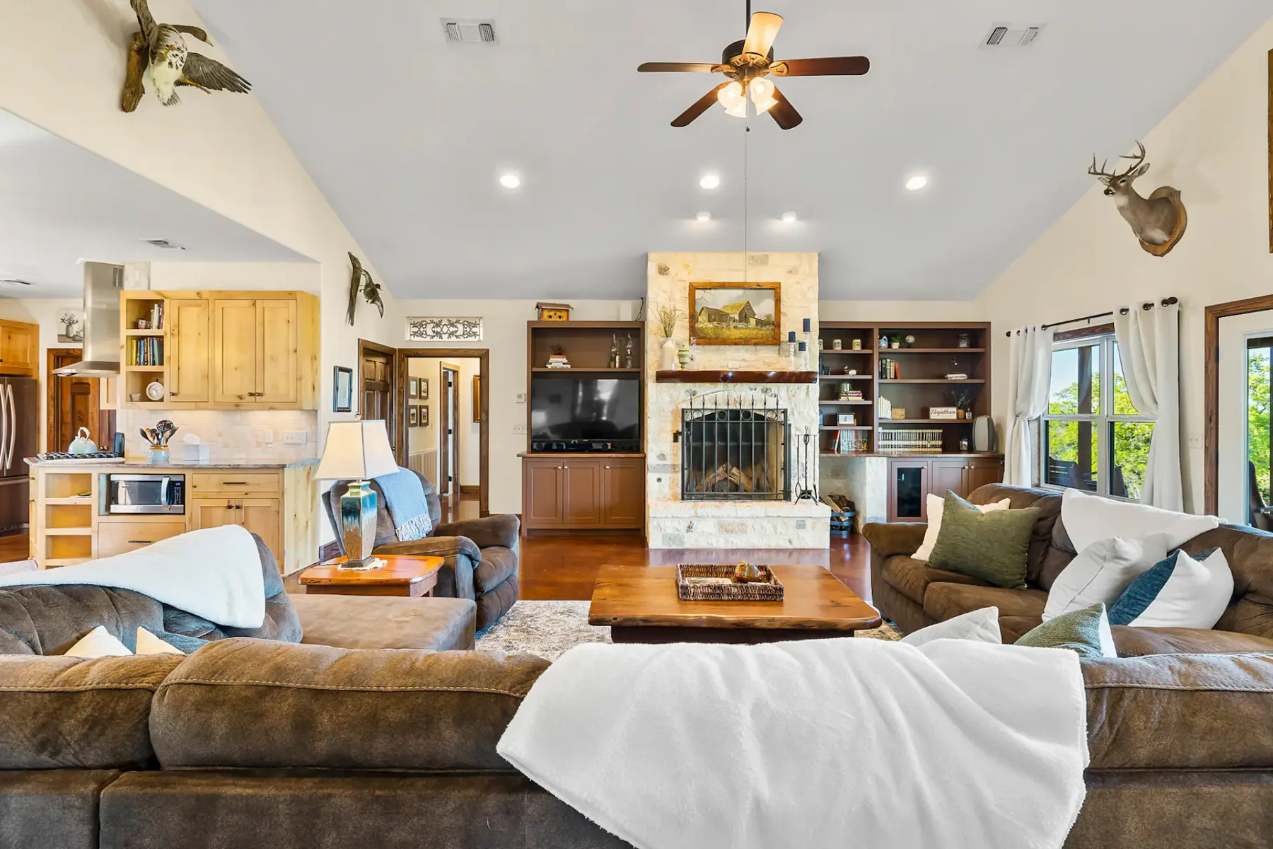 Spacious rustic living room with a stone fireplace, brown leather sofas, a ceiling fan, mounted deer heads, and wooden shelves.