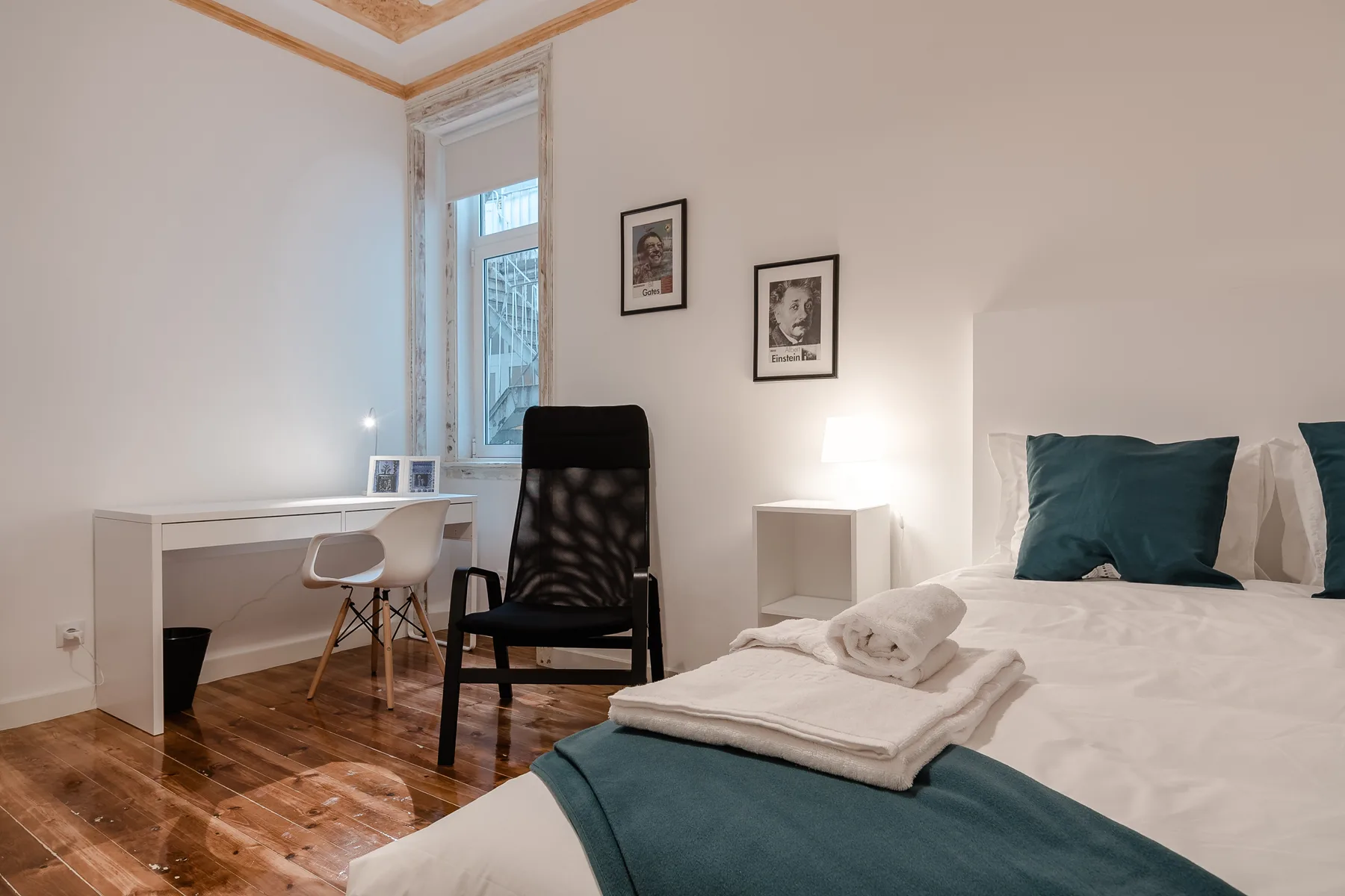 A bedroom with a bed, teal bedding, and a small desk by the window.
