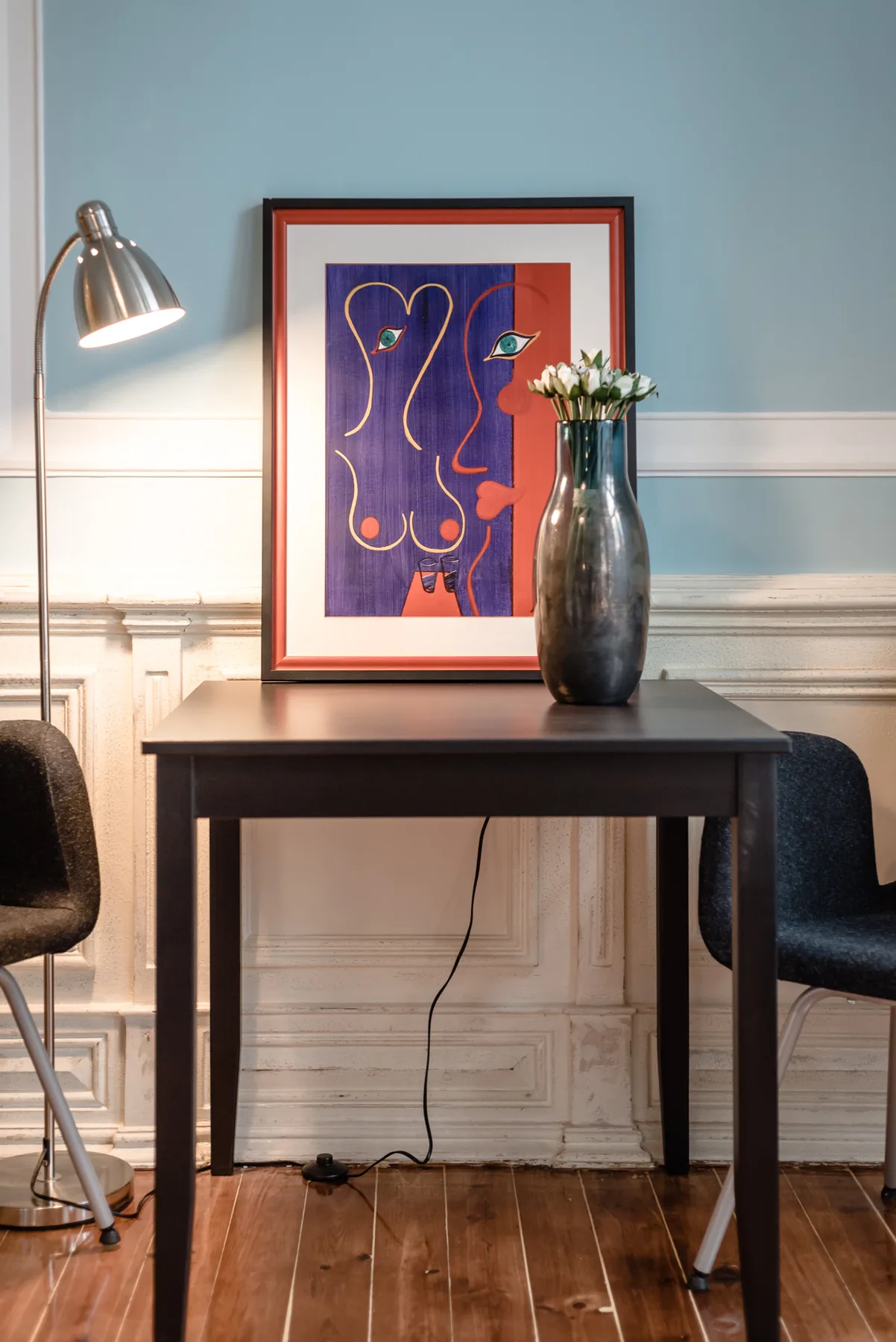 A desk sits against a blue wall, with framed art, a lamp, and a vase.
