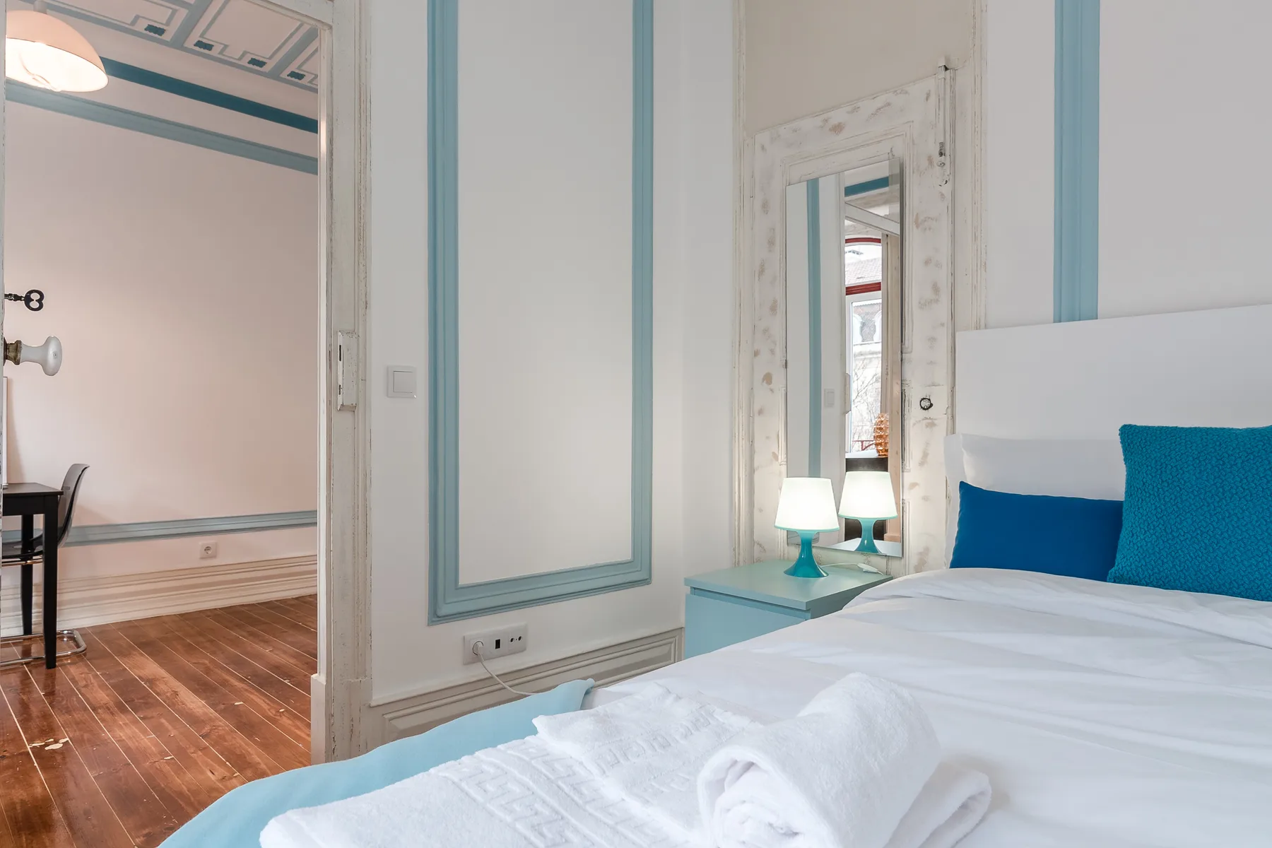 A bedroom with white bedding, blue pillows, and a bedside table.
