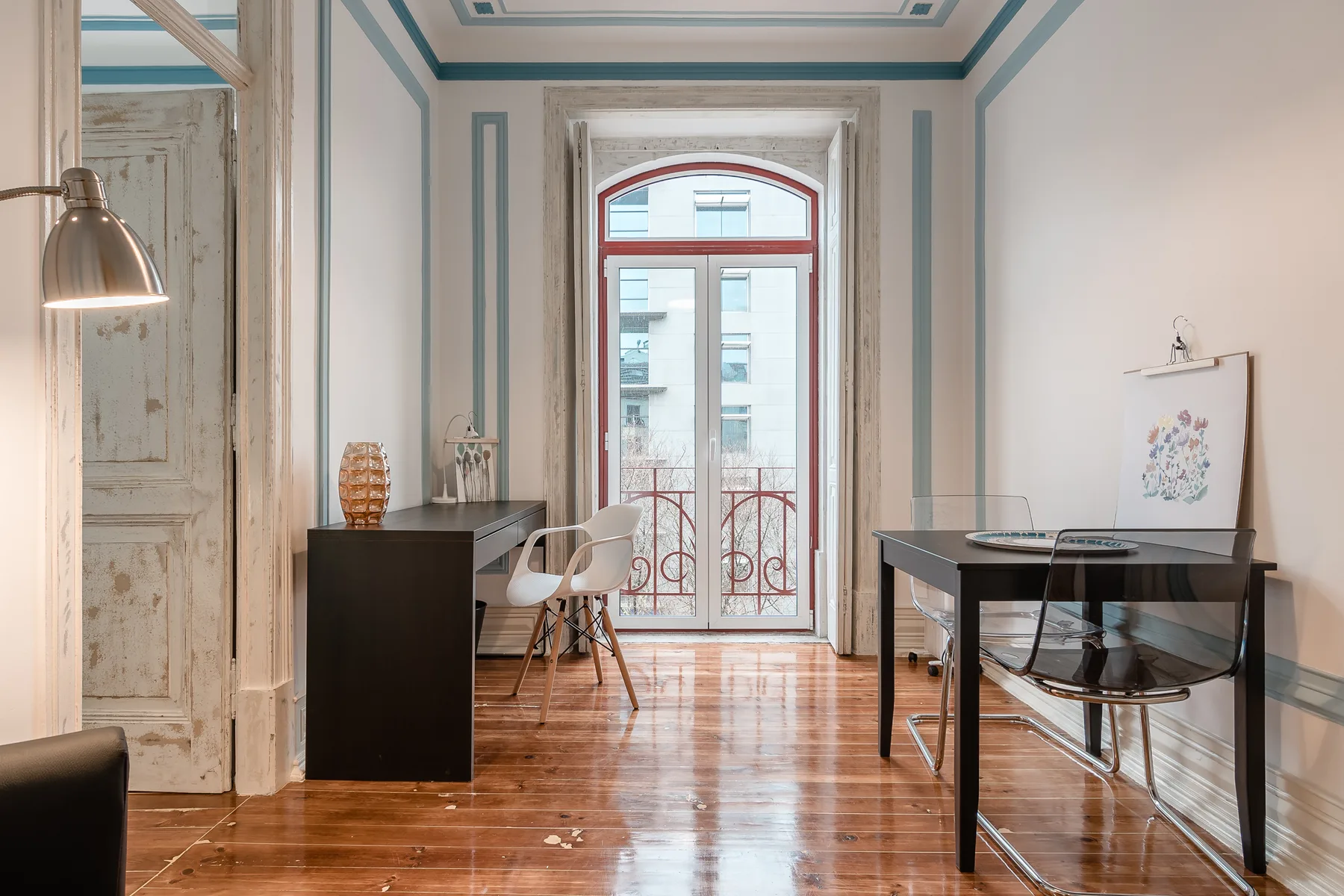 Una habitación luminosa con escritorio, asientos y una ventana alta con edificios cercanos a la vista.