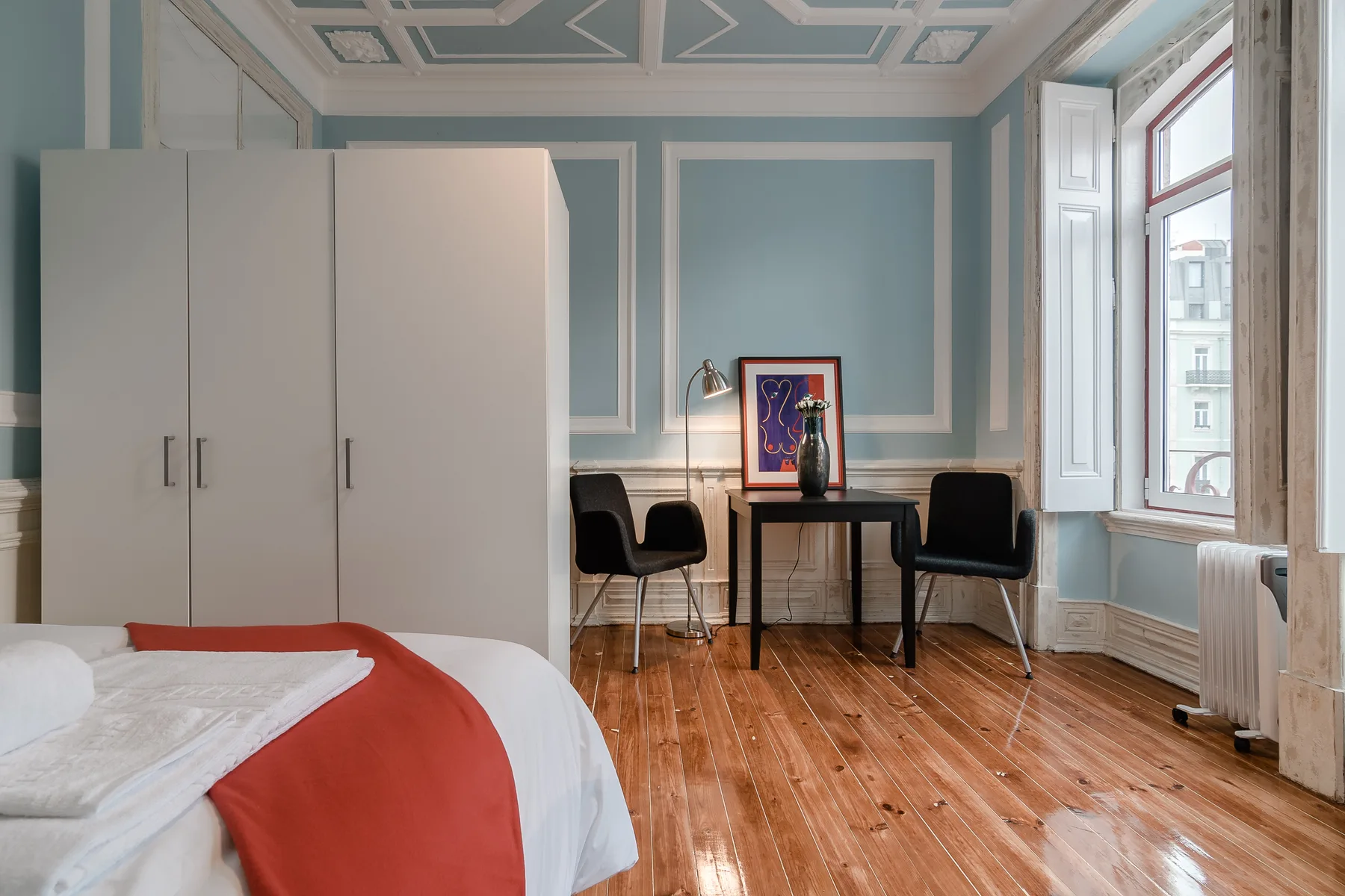A bedroom with a bed, wardrobe, desk, and chairs beside a large window.