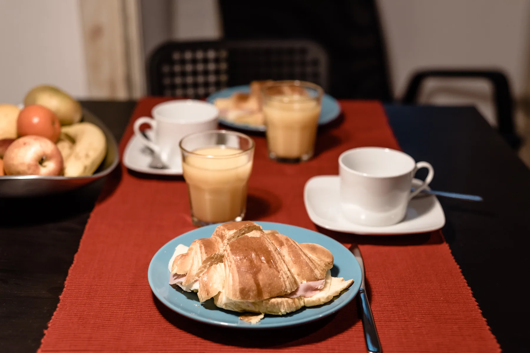 A breakfast table with croissants, coffee, cups, and a bowl of fruit.