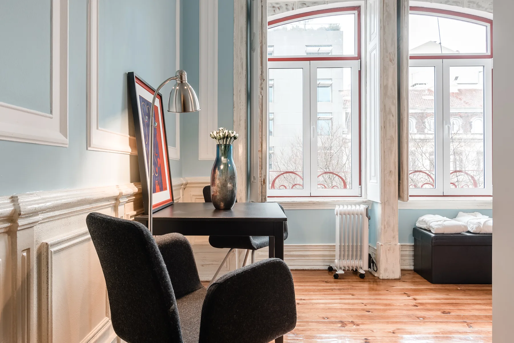 A desk and seating sit beside tall windows in a bright room.
