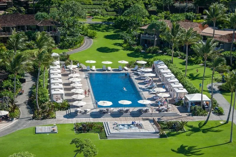 An outdoor swimming pool is surrounded by lounge chairs, umbrellas, and green landscaping.