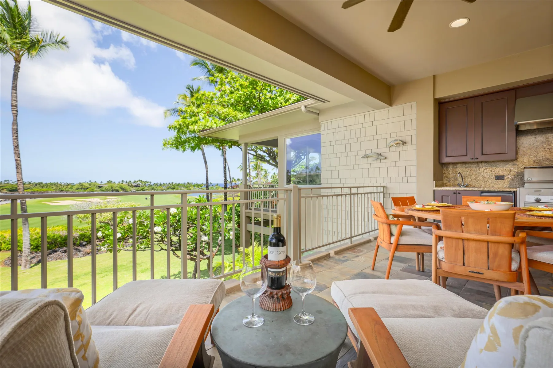 A covered outdoor dining area overlooks a green landscape, with a table and cushioned seating.