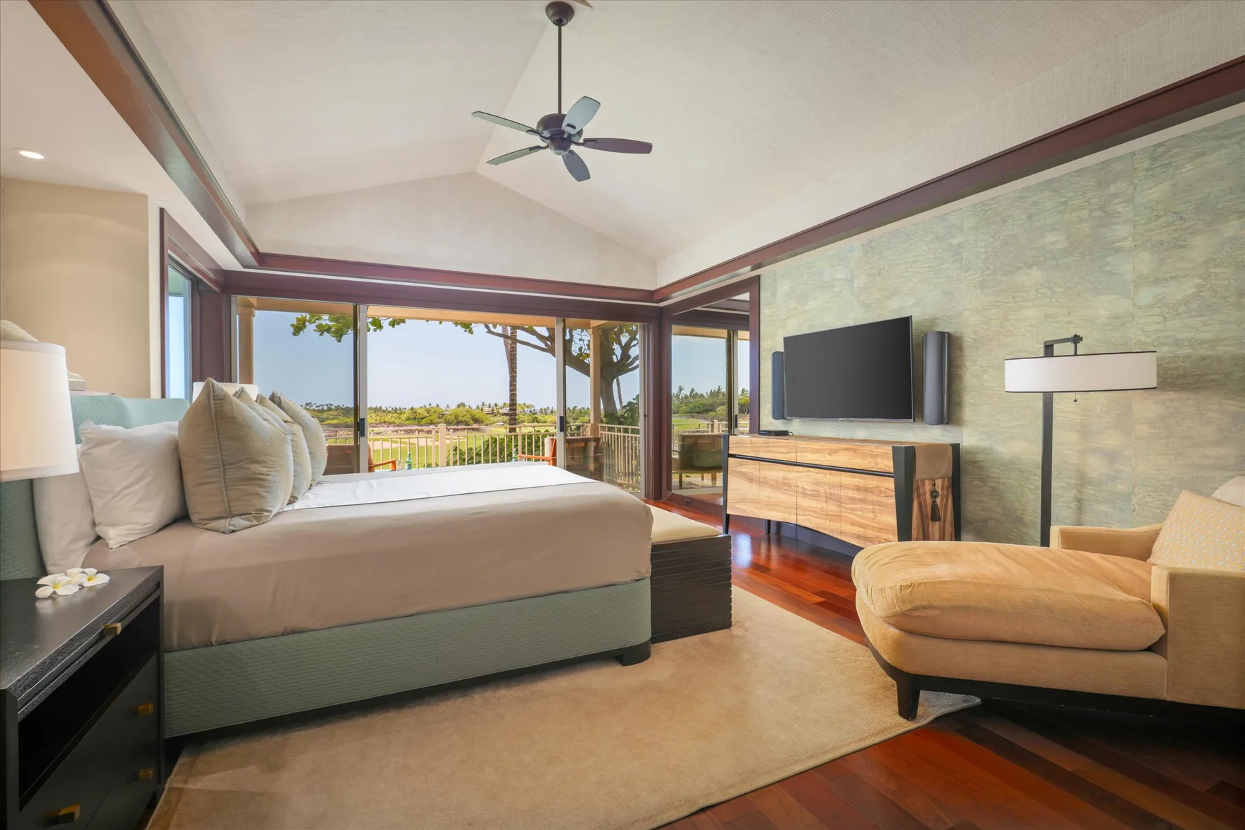 A bedroom with a large bed, a television, and windows overlooking greenery.