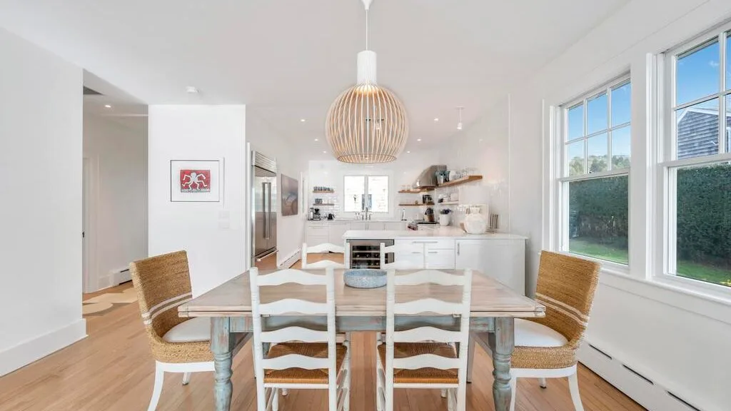 A bright dining area with a long table beside a white kitchen.
