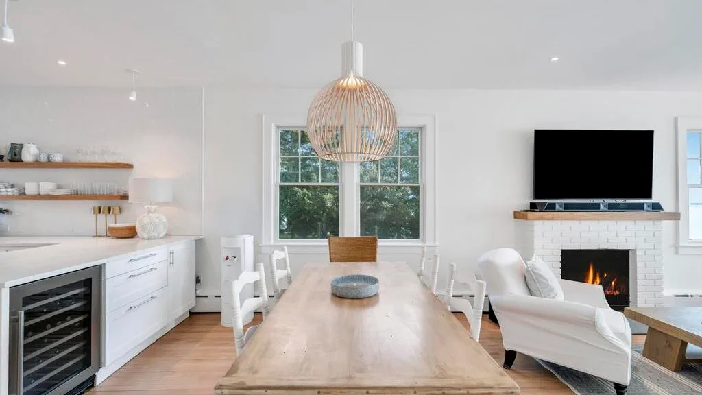A bright dining and living area with a long table, white seating, and a fireplace.
