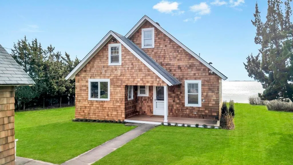 A small shingle-sided house stands beside a green lawn under a blue sky.
