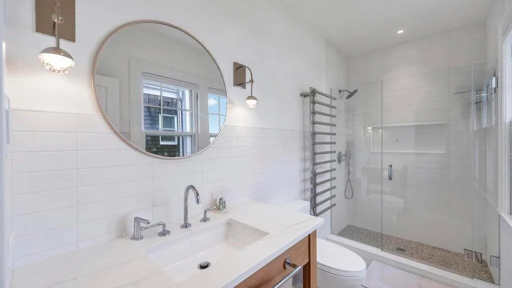 A bright bathroom has a wide sink, round mirror, and glass shower.