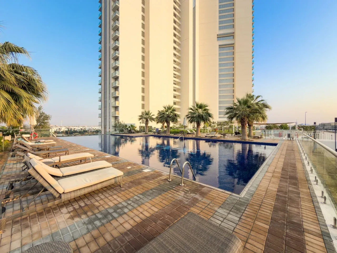 Una piscina en la azotea está rodeada de tumbonas, palmeras y un edificio alto.