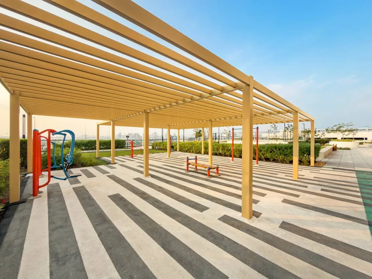 Una zona de ejercicio al aire libre está bajo una pérgola de madera, con equipos de fitness sobre el pavimento.